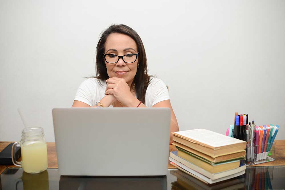 A middle-aged woman with dark brown hair and glasses is seated and looking at a laptop. A glass of lemonade is sitting on the desk to her left. To her right is a stack of books and an assortment of colorful pens.